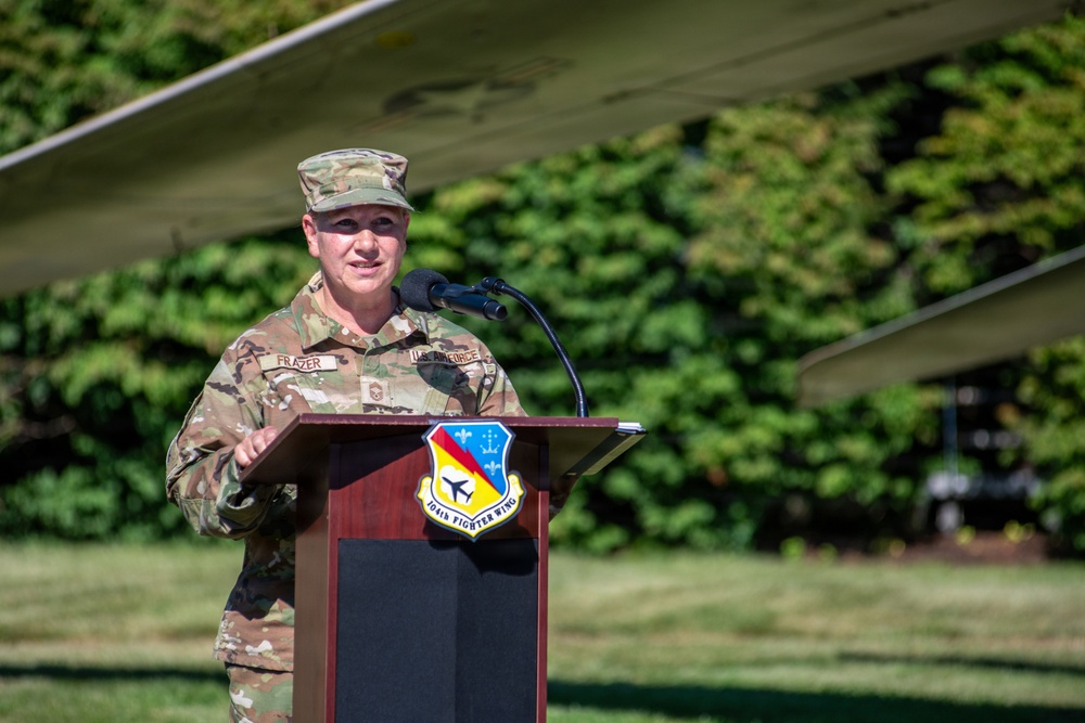 A U.S. Air Force officer with the name tape 'FRAZER' is speaking at a podium bearing the insignia of the 104th Fighter Wing. The outdoor ceremony takes place in front of what appears to be a static aircraft display, with green foliage visible in the background. The officer is wearing OCP (Operational Camouflage Pattern) uniform.