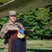 A U.S. Air Force officer with the name tape 'FRAZER' is speaking at a podium bearing the insignia of the 104th Fighter Wing. The outdoor ceremony takes place in front of what appears to be a static aircraft display, with green foliage visible in the background. The officer is wearing OCP (Operational Camouflage Pattern) uniform.