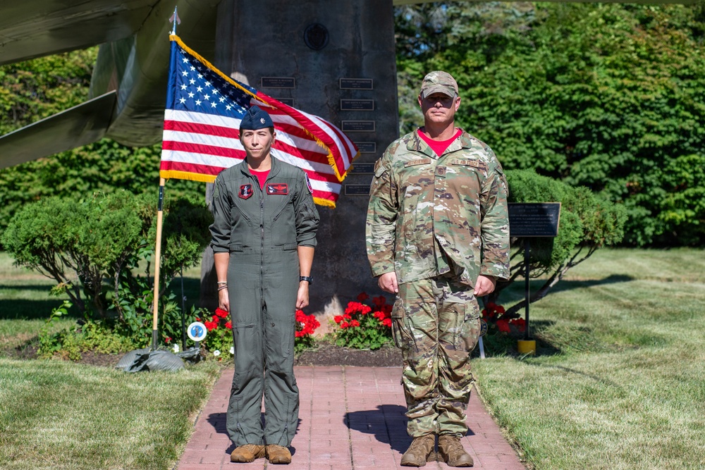 Two U.S. military personnel are standing at attention on a brick pathway in front of a memorial monument with an American flag. The individual on the left is wearing a flight suit with a garrison cap, while the individual on the right is wearing OCP (Operational Camouflage Pattern) combat uniform with a patrol cap. The setting appears to be a formal outdoor military ceremony or memorial event.