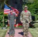 Two U.S. military personnel are standing at attention on a brick pathway in front of a memorial monument with an American flag. The individual on the left is wearing a flight suit with a garrison cap, while the individual on the right is wearing OCP (Operational Camouflage Pattern) combat uniform with a patrol cap. The setting appears to be a formal outdoor military ceremony or memorial event.