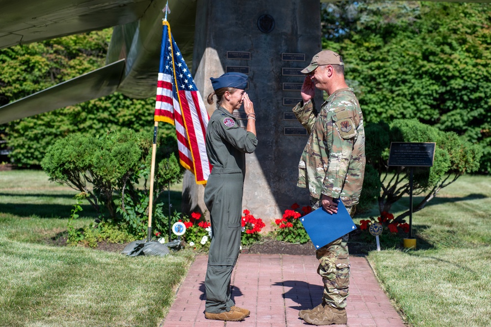 Two military personnel are exchanging salutes in an outdoor ceremonial setting. A person in an olive green flight suit and blue service cap is saluting a person in Army Combat Uniform (OCP/MultiCam) who is holding a blue folder, suggesting an official ceremony such as a promotion, award presentation, or commissioning. The setting includes an American flag, a stone memorial monument with plaques, and well-maintained grounds.
