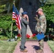 Two military personnel are exchanging salutes in an outdoor ceremonial setting. A person in an olive green flight suit and blue service cap is saluting a person in Army Combat Uniform (OCP/MultiCam) who is holding a blue folder, suggesting an official ceremony such as a promotion, award presentation, or commissioning. The setting includes an American flag, a stone memorial monument with plaques, and well-maintained grounds.
