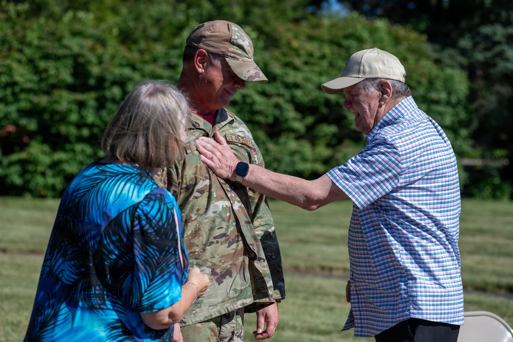An outdoor ceremony appears to be taking place involving a U.S. Air Force service member in OCP (Operational Camouflage Pattern) uniform. An older civilian man is pinning something onto the service member's chest, while a woman in a blue floral top stands nearby. The setting is a grassy outdoor area with trees in the background, suggesting a promotion or award ceremony.