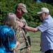 An outdoor ceremony appears to be taking place involving a U.S. Air Force service member in OCP (Operational Camouflage Pattern) uniform. An older civilian man is pinning something onto the service member's chest, while a woman in a blue floral top stands nearby. The setting is a grassy outdoor area with trees in the background, suggesting a promotion or award ceremony.