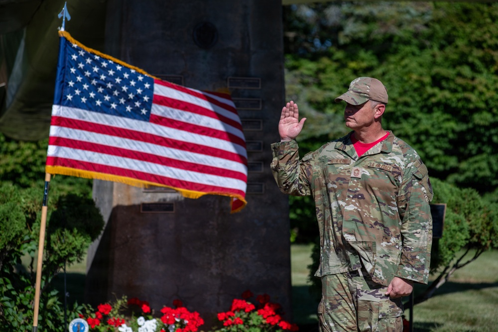 A U.S. Air Force service member in OCP (Operational Camouflage Pattern) uniform is raising their right hand, appearing to take an oath or pledge during an outdoor military ceremony. The individual stands before an American flag on a pole with gold fringe, positioned near what appears to be a memorial monument with plaques. Red, white, and blue flowers are arranged at the base of the monument, suggesting a patriotic or commemorative event.