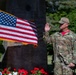 A U.S. Air Force service member in OCP (Operational Camouflage Pattern) uniform is raising their right hand, appearing to take an oath or pledge during an outdoor military ceremony. The individual stands before an American flag on a pole with gold fringe, positioned near what appears to be a memorial monument with plaques. Red, white, and blue flowers are arranged at the base of the monument, suggesting a patriotic or commemorative event.