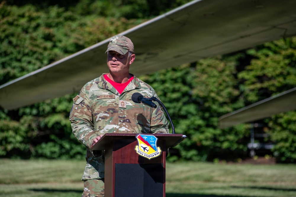 A U.S. Air Force service member wearing OCP (Operational Camouflage Pattern) uniform is speaking at a podium outdoors. The podium displays the insignia of the 104th Fighter Wing. The setting appears to be an outdoor ceremony or event, with a aircraft wing visible in the background.
