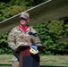 A U.S. Air Force service member wearing OCP (Operational Camouflage Pattern) uniform is speaking at a podium outdoors. The podium displays the insignia of the 104th Fighter Wing. The setting appears to be an outdoor ceremony or event, with a aircraft wing visible in the background.