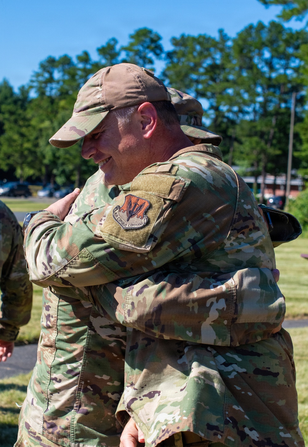 Two military personnel in OCP (Operational Camouflage Pattern) uniforms are embracing in what appears to be an outdoor ceremony or farewell event. The visible patch on one uniform identifies an Air Combat Command affiliation. The setting is a sunny, green outdoor area consistent with a military installation.