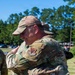 Two military personnel in OCP (Operational Camouflage Pattern) uniforms are embracing in what appears to be an outdoor ceremony or farewell event. The visible patch on one uniform identifies an Air Combat Command affiliation. The setting is a sunny, green outdoor area consistent with a military installation.