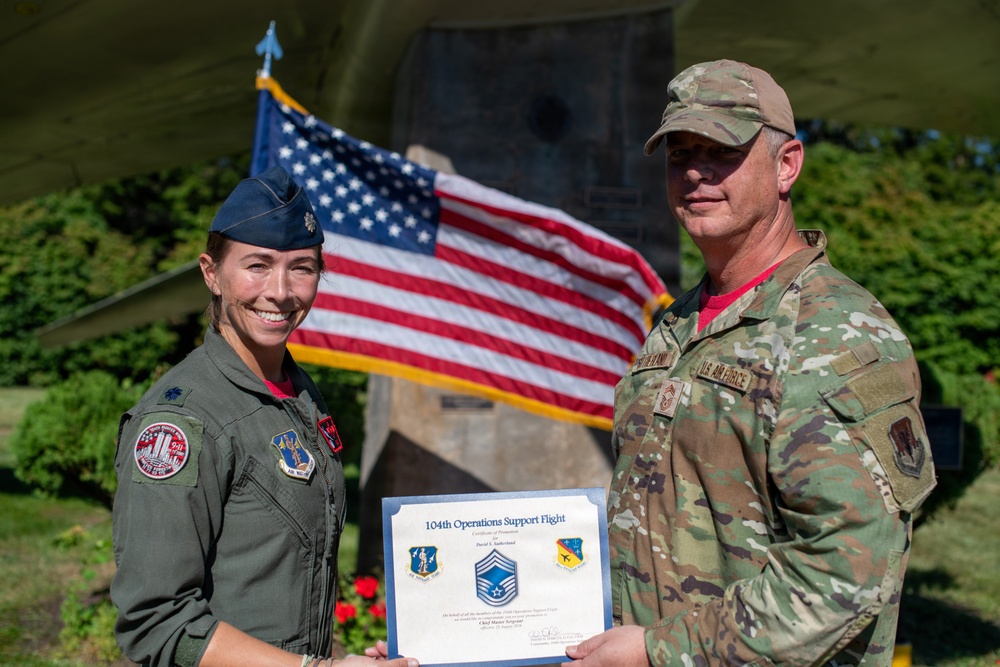 Two U.S. Air Force personnel are posing together outdoors holding a certificate from the 104th Operations Support Flight. The woman on the left is wearing an olive green flight suit with patches and a blue service cap, while the man on the right is in OCP (Operational Camouflage Pattern) uniform. An American flag is prominently displayed behind them near what appears to be a monument or memorial.