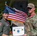 Two U.S. Air Force personnel are posing together outdoors holding a certificate from the 104th Operations Support Flight. The woman on the left is wearing an olive green flight suit with patches and a blue service cap, while the man on the right is in OCP (Operational Camouflage Pattern) uniform. An American flag is prominently displayed behind them near what appears to be a monument or memorial.