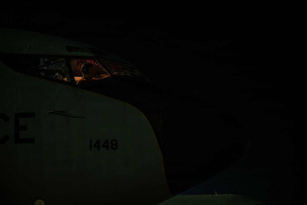 A nighttime photograph of a military aircraft cockpit with a pilot or crew member visible inside. The aircraft fuselage displays the number '1448' and partial lettering 'CE', suggesting a U.S. Air Force designation. The cockpit is dimly illuminated by red instrument lighting, creating a low-light operational atmosphere.