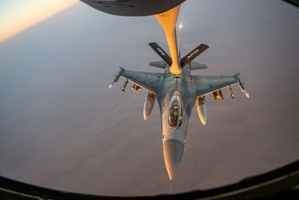 An aerial refueling operation is captured from the perspective of a tanker aircraft, showing an F-16 Fighting Falcon receiving fuel via a boom refueling system. The image is taken during golden hour, casting warm orange light across the aircraft and the arid terrain visible far below. The refueling boom is marked with the number '196' and 'ARW' (Air Refueling Wing) designation.