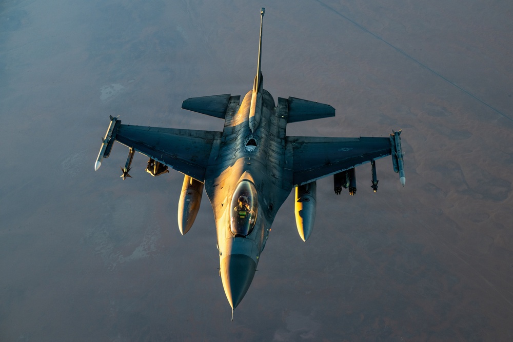 An aerial top-down photograph of a military fighter jet, consistent with an F-16 Fighting Falcon, captured in flight over an arid, desert-like terrain. The aircraft appears to be engaged in an aerial refueling operation, as indicated by the refueling boom/line visible at the top of the frame. The image is taken during golden hour lighting, casting warm amber tones across the aircraft's fuselage.