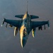 An aerial top-down photograph of a military fighter jet, consistent with an F-16 Fighting Falcon, captured in flight over an arid, desert-like terrain. The aircraft appears to be engaged in an aerial refueling operation, as indicated by the refueling boom/line visible at the top of the frame. The image is taken during golden hour lighting, casting warm amber tones across the aircraft's fuselage.