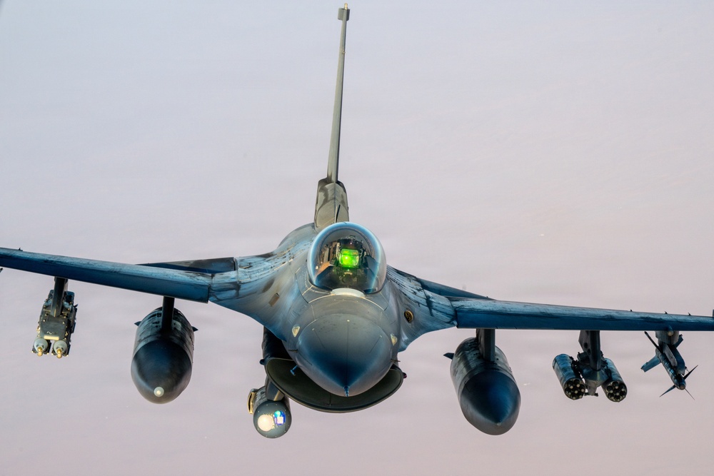A head-on photograph of a military fighter jet, consistent with an F-16 Fighting Falcon, captured in flight against an overcast sky. The aircraft is fully loaded with external ordnance and equipment on its wing pylons. The cockpit canopy is visible with a green HUD (Heads-Up Display) glow illuminated inside.