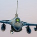 A head-on photograph of a military fighter jet, consistent with an F-16 Fighting Falcon, captured in flight against an overcast sky. The aircraft is fully loaded with external ordnance and equipment on its wing pylons. The cockpit canopy is visible with a green HUD (Heads-Up Display) glow illuminated inside.