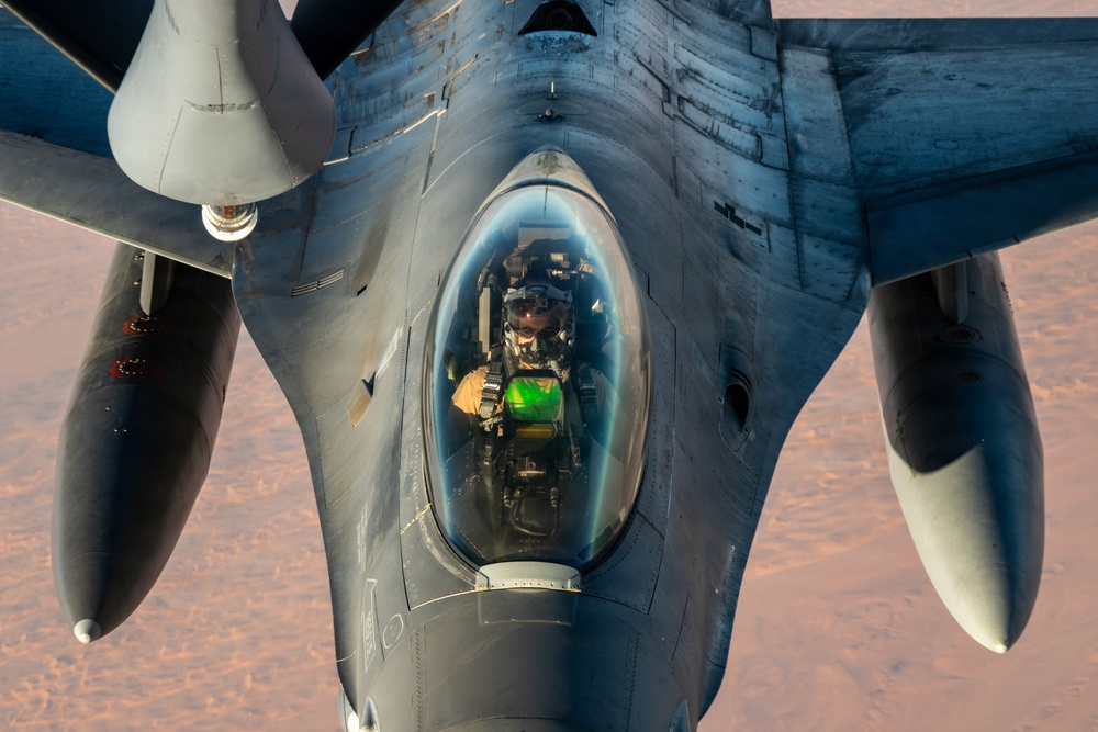 A close-up aerial photograph taken from above and behind a tanker aircraft showing an F-16 Fighting Falcon during an aerial refueling operation. The pilot is clearly visible through the bubble canopy, wearing a flight helmet and oxygen mask, with a green HUD (Heads-Up Display) glow visible in the cockpit. The aircraft is flying over what appears to be an arid desert terrain at low sun angle.