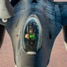A close-up aerial photograph taken from above and behind a tanker aircraft showing an F-16 Fighting Falcon during an aerial refueling operation. The pilot is clearly visible through the bubble canopy, wearing a flight helmet and oxygen mask, with a green HUD (Heads-Up Display) glow visible in the cockpit. The aircraft is flying over what appears to be an arid desert terrain at low sun angle.