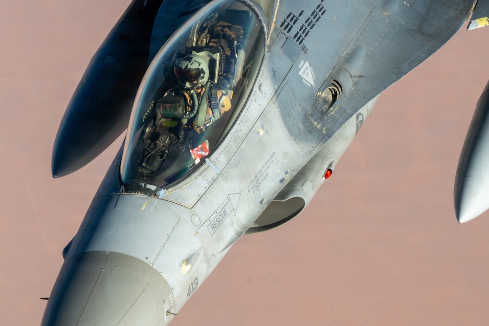 A close-up aerial photograph of an F-16 Fighting Falcon fighter jet in flight, showing the cockpit area with a pilot wearing full flight gear including helmet and oxygen mask. The aircraft bears the tail number '413' and standard military markings including a 'RESCUE' placard near the cockpit. The background suggests the aircraft is flying at altitude over a desert or arid terrain.