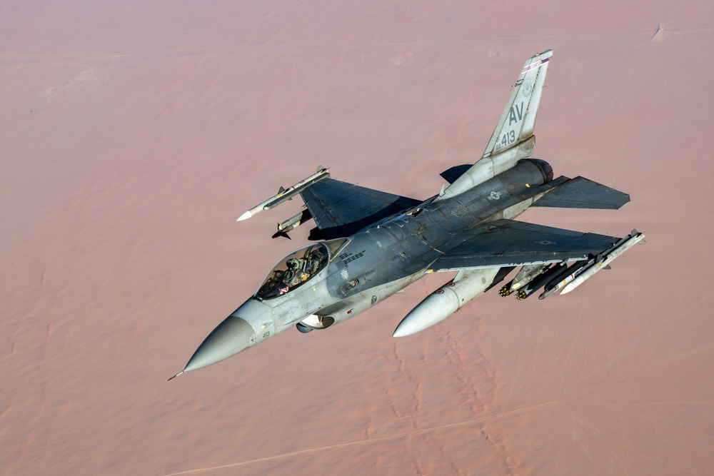An F-16 Fighting Falcon jet fighter is photographed in flight from an aerial perspective, likely from a tanker or accompanying aircraft. The aircraft bears tail code 'AV' and serial number '413', indicating it belongs to the 31st Fighter Wing based at Aviano Air Base, Italy. The aircraft is flying over an arid desert landscape and is armed with multiple air-to-air and air-to-ground munitions.