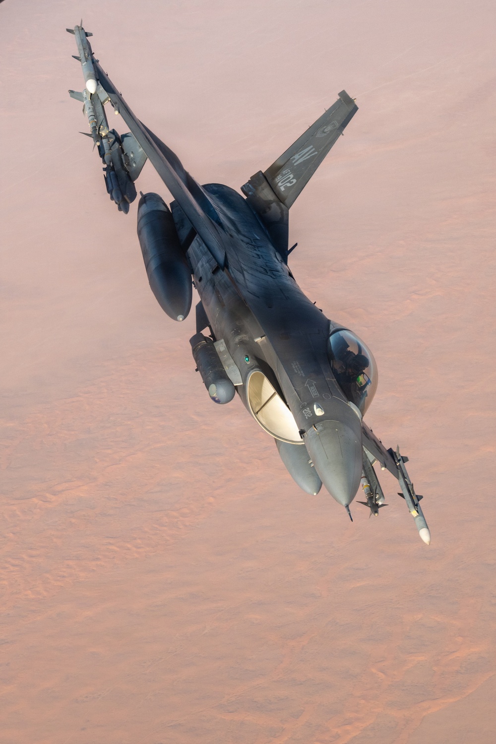 An F-16 Fighting Falcon jet fighter is photographed from above at an oblique angle while in flight over an arid, desert terrain. The aircraft is heavily armed with multiple munitions and external stores visible on its hardpoints. The tail markings and serial number are partially visible on the vertical stabilizer.