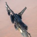 An F-16 Fighting Falcon jet fighter is photographed from above at an oblique angle while in flight over an arid, desert terrain. The aircraft is heavily armed with multiple munitions and external stores visible on its hardpoints. The tail markings and serial number are partially visible on the vertical stabilizer.
