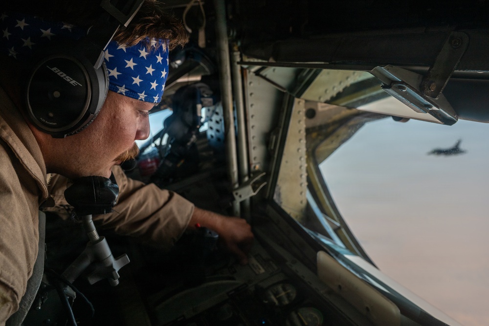 A military aircrew member wearing Bose headphones and a blue American flag-pattern bandana is seated at a station inside a large military aircraft, looking out through a window. Through the window, another military jet aircraft is visible flying in formation against a desert or arid terrain background. The individual appears to be monitoring or operating controls at their station.