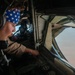 A military aircrew member wearing Bose headphones and a blue American flag-pattern bandana is seated at a station inside a large military aircraft, looking out through a window. Through the window, another military jet aircraft is visible flying in formation against a desert or arid terrain background. The individual appears to be monitoring or operating controls at their station.
