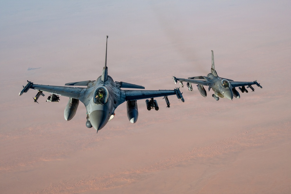 Two F-16 Fighting Falcon jet fighters are flying in formation at altitude over what appears to be an arid desert landscape. Both aircraft are armed with multiple munitions and external fuel tanks visible on their hardpoints. The image is taken from a forward-facing perspective, likely from a tanker or third aircraft flying ahead of the formation.