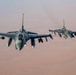 Two F-16 Fighting Falcon jet fighters are flying in formation at altitude over what appears to be an arid desert landscape. Both aircraft are armed with multiple munitions and external fuel tanks visible on their hardpoints. The image is taken from a forward-facing perspective, likely from a tanker or third aircraft flying ahead of the formation.