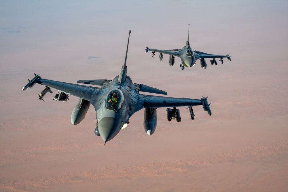 Two F-16 Fighting Falcon jet fighters are flying in formation over an arid desert landscape at what appears to be dusk or dawn, given the warm reddish-brown hues of the terrain below. Both aircraft are armed with multiple munitions and external stores on their wing pylons. A pilot is visible in the cockpit of the foreground aircraft.