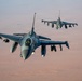 Two F-16 Fighting Falcon jet fighters are flying in formation over an arid desert landscape at what appears to be dusk or dawn, given the warm reddish-brown hues of the terrain below. Both aircraft are armed with multiple munitions and external stores on their wing pylons. A pilot is visible in the cockpit of the foreground aircraft.
