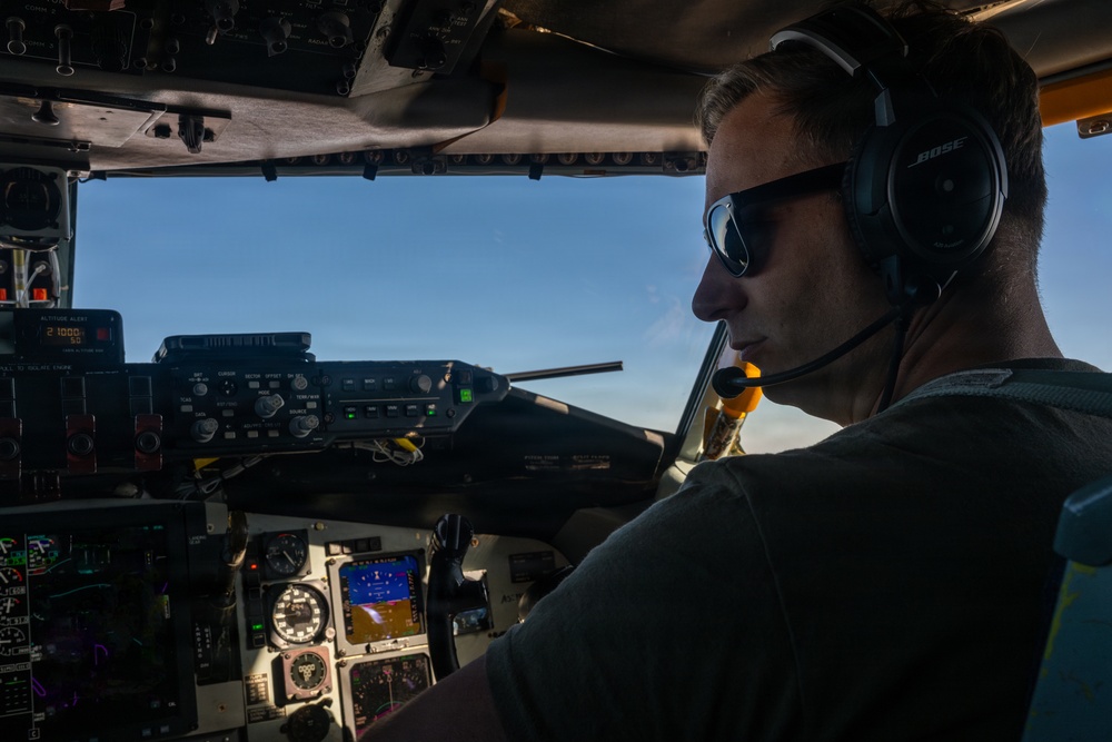 A pilot is seated in the cockpit of an aircraft in flight, wearing a Bose aviation headset with microphone and dark sunglasses. The cockpit displays multiple avionics instruments including what appears to be a glass cockpit display and an altitude alert reading approximately 21,000 feet. The image is taken from the co-pilot seat perspective with clear blue sky visible through the windscreen.