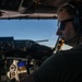 A pilot is seated in the cockpit of an aircraft in flight, wearing a Bose aviation headset with microphone and dark sunglasses. The cockpit displays multiple avionics instruments including what appears to be a glass cockpit display and an altitude alert reading approximately 21,000 feet. The image is taken from the co-pilot seat perspective with clear blue sky visible through the windscreen.