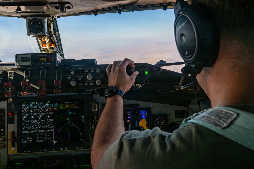 A military pilot is seated in the cockpit of a large aircraft, operating flight controls while in flight over an arid, desert-like terrain. The cockpit features a mix of analog gauges and modern digital displays, consistent with a military transport aircraft. The pilot is wearing Bose aviation headphones and a military-style flight suit.