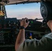 A military pilot is seated in the cockpit of a large aircraft, operating flight controls while in flight over an arid, desert-like terrain. The cockpit features a mix of analog gauges and modern digital displays, consistent with a military transport aircraft. The pilot is wearing Bose aviation headphones and a military-style flight suit.