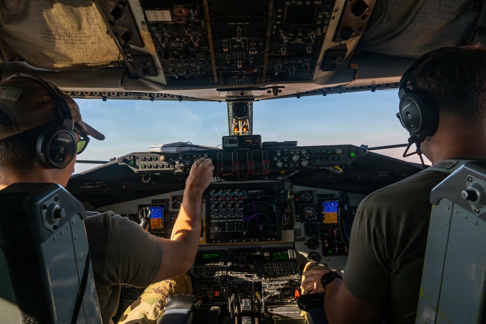 Two pilots are seated in the cockpit of a large military aircraft, likely a C-17 Globemaster III or similar transport aircraft, during flight. One pilot on the left is wearing a camouflage uniform and a cap, while the pilot on the right wears an olive green shirt. Both are equipped with Bose aviation headsets and are surrounded by an extensive array of analog and digital flight instruments.