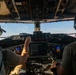 Two pilots are seated in the cockpit of a large military aircraft, likely a C-17 Globemaster III or similar transport aircraft, during flight. One pilot on the left is wearing a camouflage uniform and a cap, while the pilot on the right wears an olive green shirt. Both are equipped with Bose aviation headsets and are surrounded by an extensive array of analog and digital flight instruments.