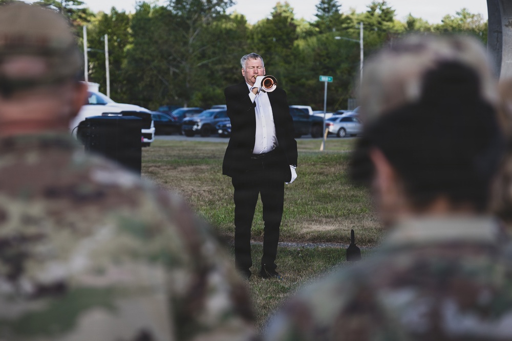 A civilian musician in formal black attire plays a bugle or trumpet outdoors while military personnel in camouflage uniforms stand in the foreground observing. The setting appears to be a military ceremony, likely a memorial or honors event such as Taps being played. The scene takes place on a grassy area with a parking lot and trees visible in the background.