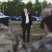 A civilian musician in formal black attire plays a bugle or trumpet outdoors while military personnel in camouflage uniforms stand in the foreground observing. The setting appears to be a military ceremony, likely a memorial or honors event such as Taps being played. The scene takes place on a grassy area with a parking lot and trees visible in the background.