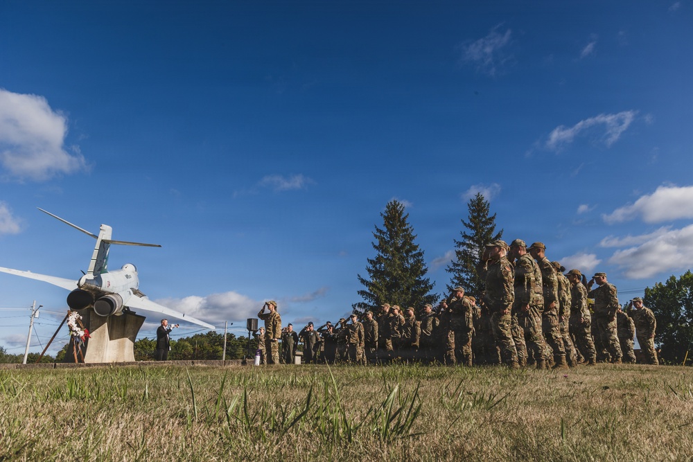 A military ceremony is taking place outdoors near a static aircraft display monument, with a wreath visible at its base. A formation of soldiers in camouflage uniforms are rendering a salute toward the monument while a civilian or official figure stands at a podium or speaking position beside it. The scene appears to be a memorial or commemorative event conducted on a grassy field under clear blue skies.