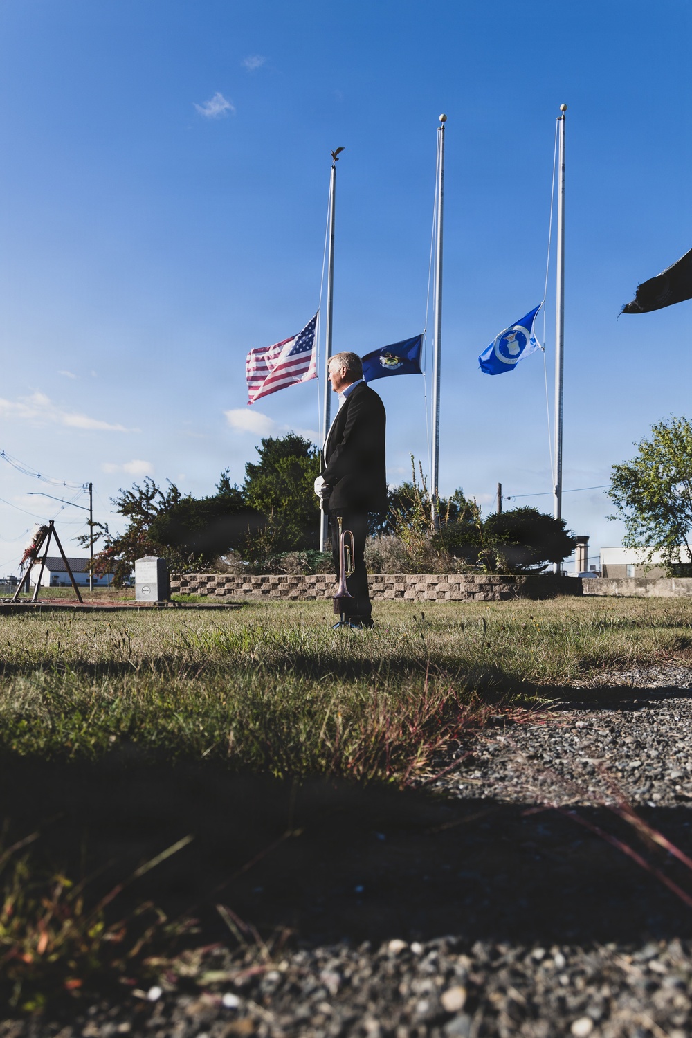 A man in formal dark attire stands at attention on a grass lawn holding a bugle at his side, positioned in front of three flagpoles flying flags at half-staff. The scene appears to be a military or civic memorial ceremony, with a small monument or marker visible to the left. The low camera angle emphasizes the solemnity of the moment against a clear blue sky.