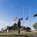 A man in formal dark attire stands at attention on a grass lawn holding a bugle at his side, positioned in front of three flagpoles flying flags at half-staff. The scene appears to be a military or civic memorial ceremony, with a small monument or marker visible to the left. The low camera angle emphasizes the solemnity of the moment against a clear blue sky.