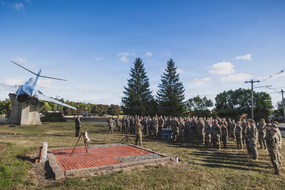 A military ceremony is taking place outdoors on what appears to be a military base or installation. A large formation of uniformed personnel in camouflage uniforms is assembled near a memorial brick plaza, with a static display aircraft (jet) mounted on a pedestal visible to the left. A civilian or official in dark clothing appears to be addressing the assembled troops.