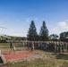 A military ceremony is taking place outdoors on what appears to be a military base or installation. A large formation of uniformed personnel in camouflage uniforms is assembled near a memorial brick plaza, with a static display aircraft (jet) mounted on a pedestal visible to the left. A civilian or official in dark clothing appears to be addressing the assembled troops.