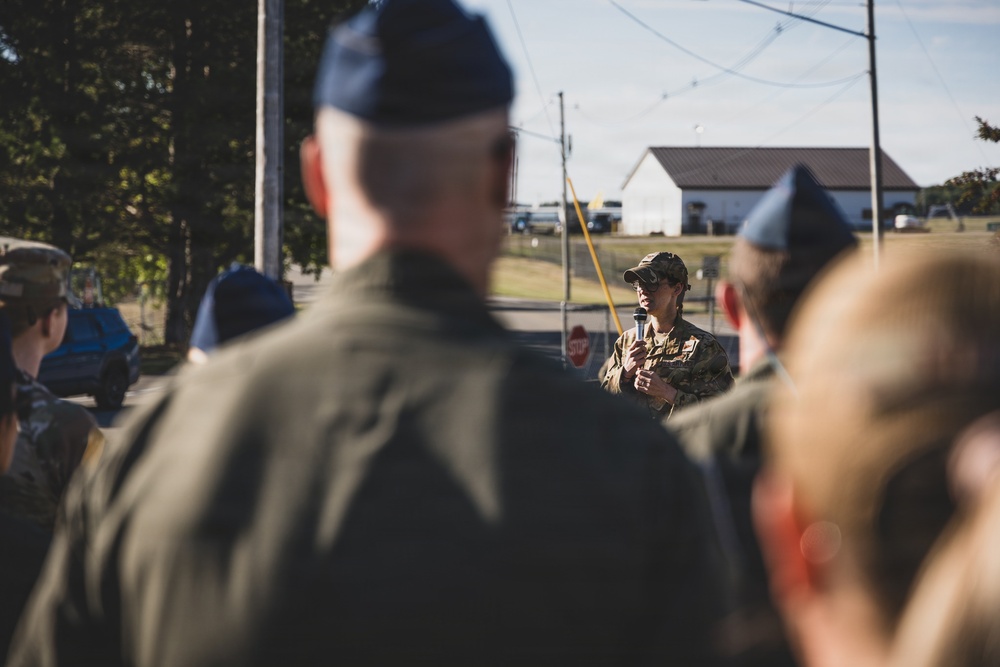 A military personnel in camouflage uniform and cap is addressing a group of service members outdoors, holding a microphone. The audience, visible from behind, appears to be wearing dark flight suits and blue garrison caps, consistent with U.S. Air Force dress. The setting appears to be a military installation or airfield area during daylight hours.