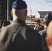 A military personnel in camouflage uniform and cap is addressing a group of service members outdoors, holding a microphone. The audience, visible from behind, appears to be wearing dark flight suits and blue garrison caps, consistent with U.S. Air Force dress. The setting appears to be a military installation or airfield area during daylight hours.