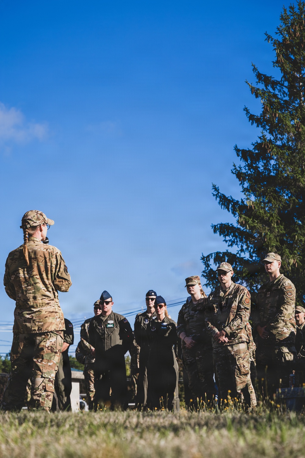 A military briefing or instruction session taking place outdoors on a grassy field under a clear blue sky. One individual in OCP (Operational Camouflage Pattern) uniform faces a group of approximately 8-10 military personnel, some in OCP uniforms and others in dark flight suits. The setting and attire suggest a U.S. Air Force training or operational exercise.